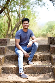 A man is sitting on outdoor stone steps surrounded by a natural setting with trees and greenery in the background. He is wearing a blue t-shirt and jeans, and smiling while looking at the camera.