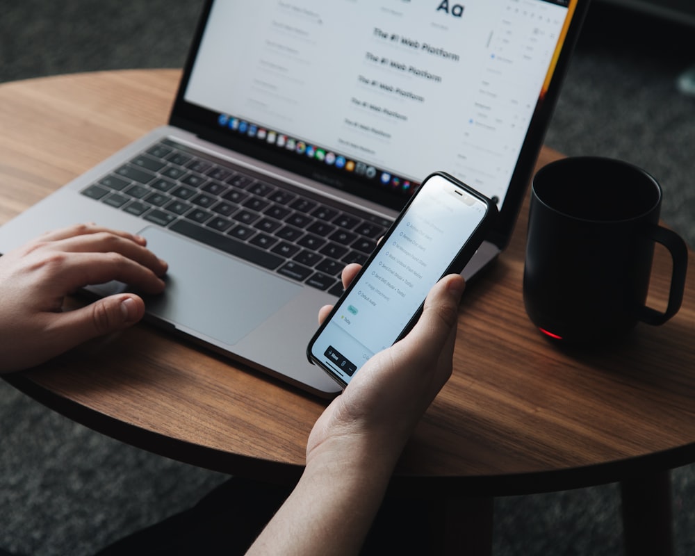 person holding smartphone in front of turned-on laptop beside black ceramic mug on oval brown wooden table