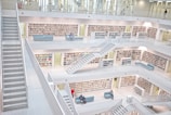 A modern library with white interior and multiple floors featuring open bookshelves filled with books. There are several staircases connecting the levels and seating areas with people reading or using electronic devices.