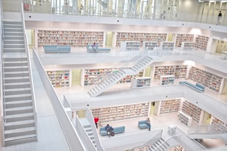 A modern library with white interior and multiple floors featuring open bookshelves filled with books. There are several staircases connecting the levels and seating areas with people reading or using electronic devices.