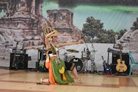 A dancer in traditional costume performs on stage, featuring vibrant colors and intricate patterns. The backdrop includes ancient ruins and lush greenery. Several musical instruments, such as guitars and drums, are visible, suggesting a cultural performance.