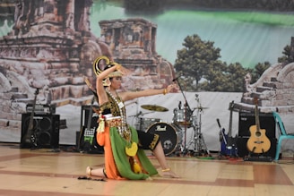 A dancer in traditional costume performs on stage, featuring vibrant colors and intricate patterns. The backdrop includes ancient ruins and lush greenery. Several musical instruments, such as guitars and drums, are visible, suggesting a cultural performance.