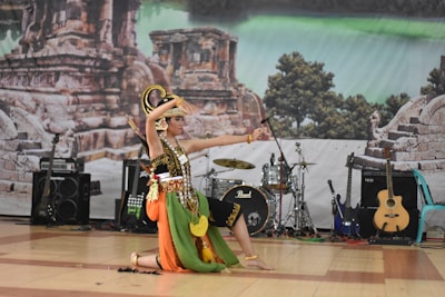A dancer in traditional costume performs on stage, featuring vibrant colors and intricate patterns. The backdrop includes ancient ruins and lush greenery. Several musical instruments, such as guitars and drums, are visible, suggesting a cultural performance.
