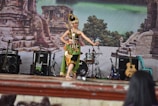 A performer dressed in traditional attire is dancing on a stage. The stage is set against a backdrop depicting ancient stone ruins and lush greenery. There are several musical instruments around, including guitars, an amplifier, and a drum set. Several chairs can also be seen near the stage.