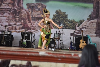 A performer dressed in traditional attire is dancing on a stage. The stage is set against a backdrop depicting ancient stone ruins and lush greenery. There are several musical instruments around, including guitars, an amplifier, and a drum set. Several chairs can also be seen near the stage.