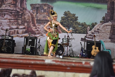 A performer dressed in traditional attire is dancing on a stage. The stage is set against a backdrop depicting ancient stone ruins and lush greenery. There are several musical instruments around, including guitars, an amplifier, and a drum set. Several chairs can also be seen near the stage.