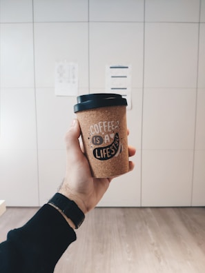 A hand holding a coffee cup with an inscription, set against a minimalist, light-colored background. The cup has a textured design with the phrase 'Coffee is a lifestyle' printed on it. The person wearing a black wristband or watch is visible.