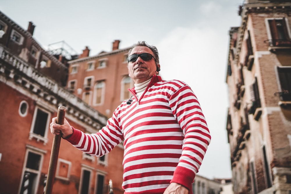 man in white and red stripe long-sleeve shirt holding stick while standing near building under white sky during day time