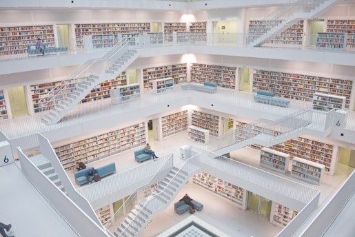 A modern, multi-level library interior with white walls and numerous bookshelves filled with colorful books. Several people are sitting on blue couches scattered around the open space. Multiple staircases connect the different levels.