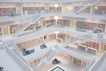 A modern, multi-level library interior with white walls and numerous bookshelves filled with colorful books. Several people are sitting on blue couches scattered around the open space. Multiple staircases connect the different levels.