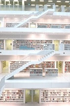 A modern, multi-level library interior with a clean, minimalist design. The space features open bookshelves filled with colorful books and several levels connected by white staircases. The environment is well-lit with a spacious, airy feel, creating an atmosphere conducive to reading and study.
