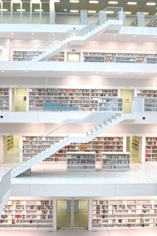 A modern, multi-level library interior with a clean, minimalist design. The space features open bookshelves filled with colorful books and several levels connected by white staircases. The environment is well-lit with a spacious, airy feel, creating an atmosphere conducive to reading and study.
