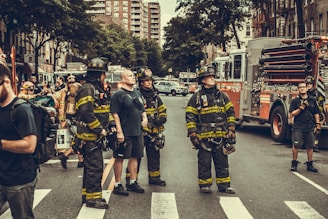 A group of firefighters stand in the street, wearing full gear with helmets and protective clothing. Several fire trucks and emergency vehicles are visible in the background. The scene is set in an urban environment, with buildings lining the street and a crowd of people, possibly part of the emergency response team, gathered around.