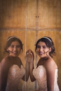 A young woman in an elegant, floral-patterned gown is standing beside a mirrored surface with her reflection visible. Her hair is styled with a delicate tiara, and she is smiling brightly, showing braces.