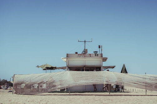 A small kiosk is situated on a sandy beach, surrounded by a large net that stretches across the foreground. The structure features a simple, rectangular design with a few antennas on its roof. An umbrella is perched to the left of the kiosk, providing shade. The sky above is clear and blue, while the sand is golden and expansive.