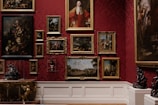 A richly decorated gallery wall features a collection of classical paintings, each in elaborate gold frames. The wall is adorned with a deep red damask pattern, adding to the opulence of the room. The artwork varies from portraits to landscapes and still life compositions. Intricately designed sculptures and ornate furniture add to the luxurious atmosphere.