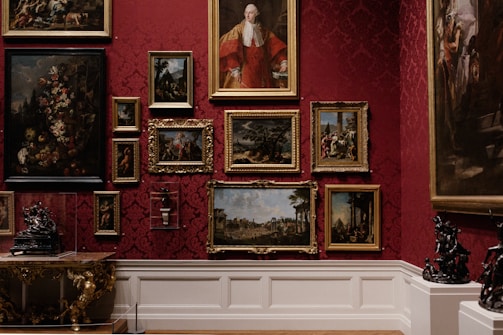A richly decorated gallery wall features a collection of classical paintings, each in elaborate gold frames. The wall is adorned with a deep red damask pattern, adding to the opulence of the room. The artwork varies from portraits to landscapes and still life compositions. Intricately designed sculptures and ornate furniture add to the luxurious atmosphere.