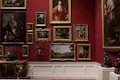 A richly decorated gallery wall features a collection of classical paintings, each in elaborate gold frames. The wall is adorned with a deep red damask pattern, adding to the opulence of the room. The artwork varies from portraits to landscapes and still life compositions. Intricately designed sculptures and ornate furniture add to the luxurious atmosphere.