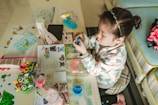 A child sits at a table filled with arts and crafts materials, including colored paper, markers, and a bunny-shaped object. The child is focused on shaping a blue dough-like material with their hands. The table is cluttered with various colorful items, including educational papers and a water bottle.