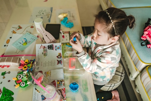 A child sits at a table filled with arts and crafts materials, including colored paper, markers, and a bunny-shaped object. The child is focused on shaping a blue dough-like material with their hands. The table is cluttered with various colorful items, including educational papers and a water bottle.