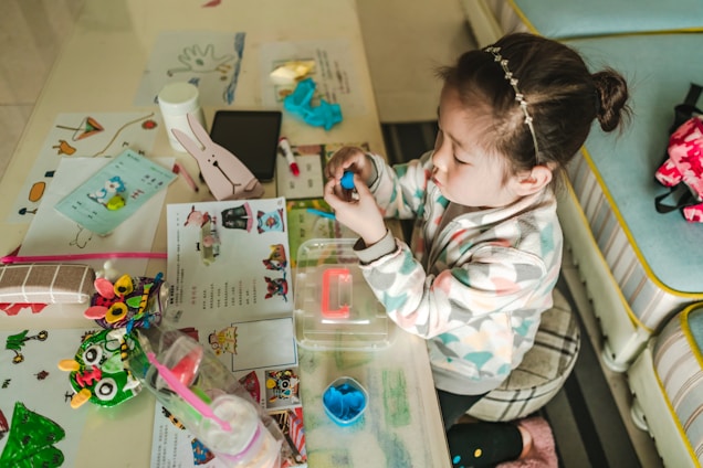 A child sits at a table filled with arts and crafts materials, including colored paper, markers, and a bunny-shaped object. The child is focused on shaping a blue dough-like material with their hands. The table is cluttered with various colorful items, including educational papers and a water bottle.