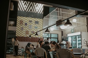 A well-lit cafe interior features a group of people sitting at a table, engaging in conversation. The atmosphere is cozy with eclectic, patterned decor on the walls, including bold chevrons and various prints. Several light fixtures hang from the ceiling, and a visible fridge indicates refreshments are available.