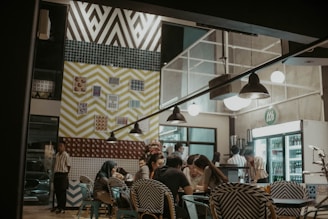 A well-lit cafe interior features a group of people sitting at a table, engaging in conversation. The atmosphere is cozy with eclectic, patterned decor on the walls, including bold chevrons and various prints. Several light fixtures hang from the ceiling, and a visible fridge indicates refreshments are available.