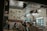 A well-lit cafe interior features a group of people sitting at a table, engaging in conversation. The atmosphere is cozy with eclectic, patterned decor on the walls, including bold chevrons and various prints. Several light fixtures hang from the ceiling, and a visible fridge indicates refreshments are available.