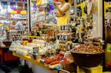 The image depicts a busy market stall filled with a variety of goods. There are numerous food items, including packaged and unpackaged products like cheeses, nuts, and bottles of liquid. The stall is well-stocked with many different kinds of products arranged neatly. A person wearing a yellow apron and white shirt is standing behind the counter, seemingly ready to assist customers. Another person can be seen in the background browsing through different items. The environment is colorful and appears to be lively.