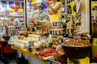 The image depicts a busy market stall filled with a variety of goods. There are numerous food items, including packaged and unpackaged products like cheeses, nuts, and bottles of liquid. The stall is well-stocked with many different kinds of products arranged neatly. A person wearing a yellow apron and white shirt is standing behind the counter, seemingly ready to assist customers. Another person can be seen in the background browsing through different items. The environment is colorful and appears to be lively.