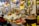 The image depicts a busy market stall filled with a variety of goods. There are numerous food items, including packaged and unpackaged products like cheeses, nuts, and bottles of liquid. The stall is well-stocked with many different kinds of products arranged neatly. A person wearing a yellow apron and white shirt is standing behind the counter, seemingly ready to assist customers. Another person can be seen in the background browsing through different items. The environment is colorful and appears to be lively.