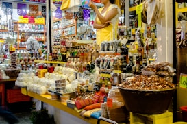 The image depicts a busy market stall filled with a variety of goods. There are numerous food items, including packaged and unpackaged products like cheeses, nuts, and bottles of liquid. The stall is well-stocked with many different kinds of products arranged neatly. A person wearing a yellow apron and white shirt is standing behind the counter, seemingly ready to assist customers. Another person can be seen in the background browsing through different items. The environment is colorful and appears to be lively.