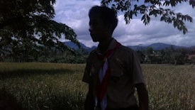 A person stands in a grassy field under the shade of a tree, with mountains visible in the distance and clouds in the sky. The individual is wearing a scout uniform, including a scarf.