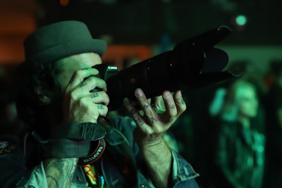 A person wearing a hat and holding a large camera with a telephoto lens, focusing intently while taking a photograph in a dimly lit environment. Tattoos are visible on the person's hands and fingers.