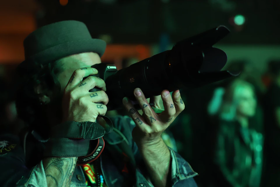 Person holding camera with both hands, wearing hat and jacket, in dimly lit room