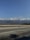 A wide open road stretches ahead, bordered by a low guardrail. In the background, a vast range of mountains is partially obscured by fluffy clouds under a clear blue sky. The shadows of nearby trees can be seen on the road surface.