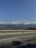 A wide open road stretches ahead, bordered by a low guardrail. In the background, a vast range of mountains is partially obscured by fluffy clouds under a clear blue sky. The shadows of nearby trees can be seen on the road surface.
