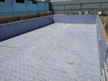 An empty swimming pool with blue and white mosaic tiles lining the interior. The surrounding area appears to be under construction with various materials scattered around. Industrial-looking walls with a corrugated metal fence are visible in the background.