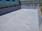 An empty swimming pool with blue and white mosaic tiles lining the interior. The surrounding area appears to be under construction with various materials scattered around. Industrial-looking walls with a corrugated metal fence are visible in the background.