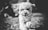 A small, fluffy dog running directly towards the camera with its mouth open, appearing playful and energetic. The background is blurred, possibly depicting grass and trees, suggesting an outdoor setting. The image is black and white, adding a classic touch.