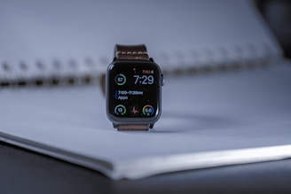 A smart watch with a digital display showing the time as 7:29, positioned on a coiled notebook. The watch face displays circular indicators for apps and health metrics. The setting is softly lit, creating a subtle contrast with the dark watch face.