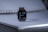 A smart watch with a digital display showing the time as 7:29, positioned on a coiled notebook. The watch face displays circular indicators for apps and health metrics. The setting is softly lit, creating a subtle contrast with the dark watch face.