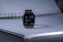 A smart watch with a digital display showing the time as 7:29, positioned on a coiled notebook. The watch face displays circular indicators for apps and health metrics. The setting is softly lit, creating a subtle contrast with the dark watch face.