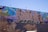 A vibrant mural covers a wall, featuring large, stylized text that appears to be an intricate combination of colors and patterns. The background includes abstract designs in shades of blue, pink, green, and yellow. The foreground shows some leafless trees and a streetlamp.