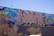 A vibrant mural covers a wall, featuring large, stylized text that appears to be an intricate combination of colors and patterns. The background includes abstract designs in shades of blue, pink, green, and yellow. The foreground shows some leafless trees and a streetlamp.