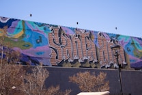 A vibrant mural covers a wall, featuring large, stylized text that appears to be an intricate combination of colors and patterns. The background includes abstract designs in shades of blue, pink, green, and yellow. The foreground shows some leafless trees and a streetlamp.