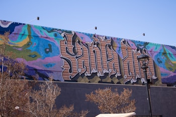 A vibrant mural covers a wall, featuring large, stylized text that appears to be an intricate combination of colors and patterns. The background includes abstract designs in shades of blue, pink, green, and yellow. The foreground shows some leafless trees and a streetlamp.