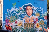 A vibrant mural on a building wall depicting a large face with expressive eyes and flowing hair decorated with various elements like hummingbirds. The hands are outstretched, surrounded by colorful flowers, swirls, and nature motifs. The background is predominantly blue, complementing the bright colors of the artwork.