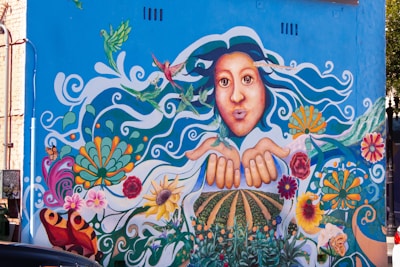A vibrant mural on a building wall depicting a large face with expressive eyes and flowing hair decorated with various elements like hummingbirds. The hands are outstretched, surrounded by colorful flowers, swirls, and nature motifs. The background is predominantly blue, complementing the bright colors of the artwork.