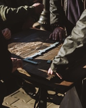 Several individuals are gathered around a wooden table engaging in a game of dominoes. The sunlight casts shadows across the table, highlighting the domino pieces meticulously arranged in play.